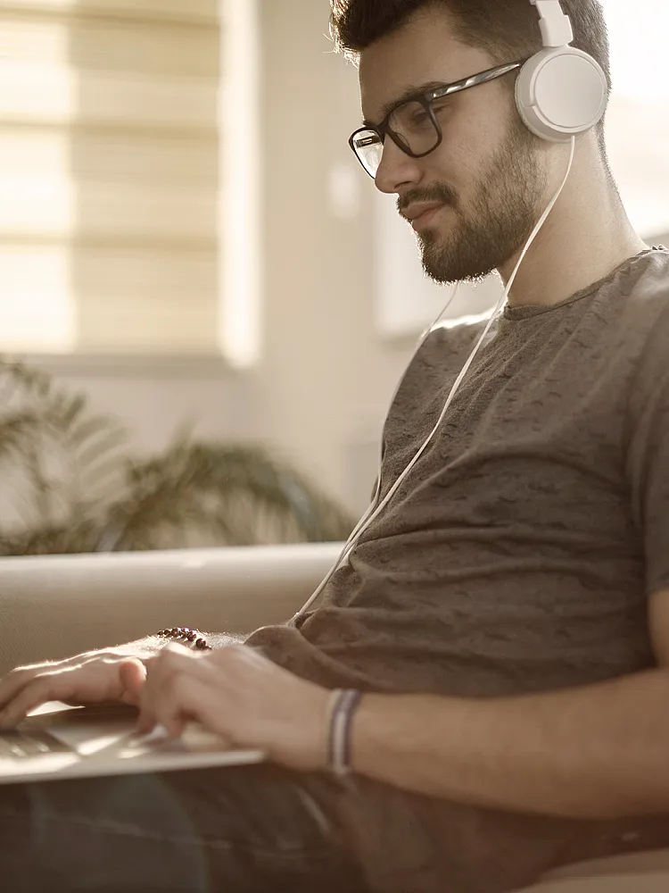 Ein junger Mann mit Brille und Tshirt trägt Kopfhörer und arbeitet an seinem Laptop