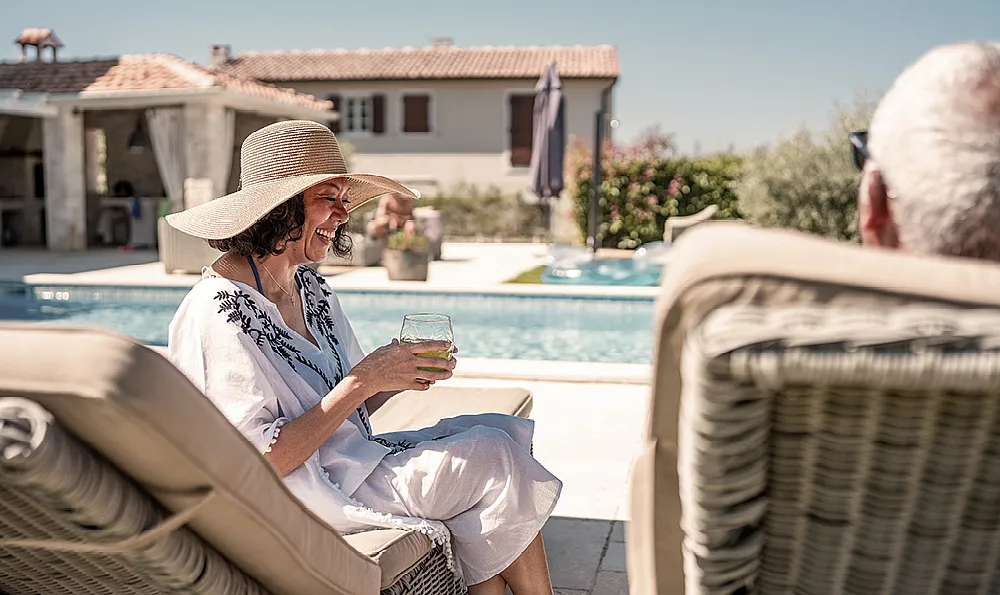 Eine Frau mit einem breitkrempigen Hut lächelt und hält ein Getränk in der Hand, während sie sich am Pool entspannt. Sonniger Tag, entspannte Atmosphäre, mit einem Steinhaus im Hintergrund.