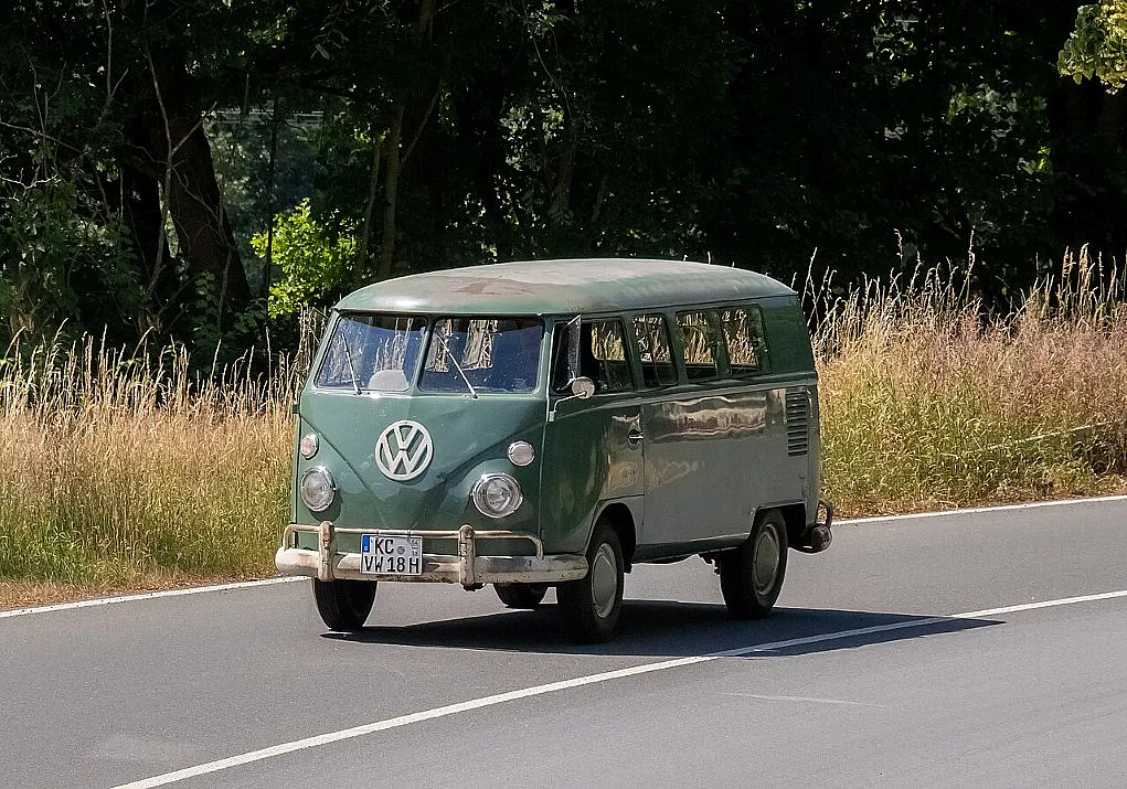 Fotografie eines grünen Volkswagen T1 Bulli, der auf einer Landstraße mit Bäumen und hohem Gras im Hintergrund fährt. Das Fahrzeug zeigt typische Merkmale wie runde Scheinwerfer, großes VW-Logo vorne und ein deutsches Kennzeichen.