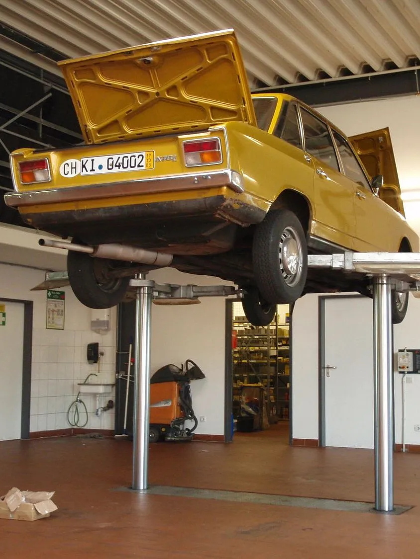 Ein gelbes Oldtimer-Auto VW K70 L 1971 steht auf einer Hebebühne in einer Garage, wobei sowohl die Motorhaube als auch der Kofferraum geöffnet sind. Die Kulisse vermittelt die Atmosphäre einer Autowerkstatt.
