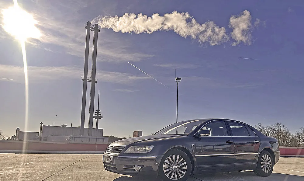 Eine schwarze VW Phaeton D1 3.0 TDI Individual 2007 parkt unter einem klaren blauen Himmel mit einer hellen Sonne in der Ecke. Im Hintergrund steigt Rauch aus hohen Industrieschornsteinen auf.
