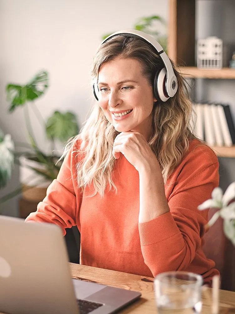 Eine Frau mit Kopfhörern und in einem orangen Pullover sitzt an einem Laptop und lächelt in die Kamera