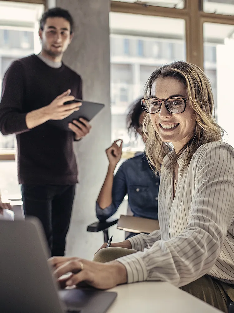 Person sitzt an einem Laptop in einem modernen Büro und arbeitet konzentriert, während zwei weitere Personen im Hintergrund stehen und auf ein Tablet schauen. Heller Raum mit großen Fenstern im Hintergrund.