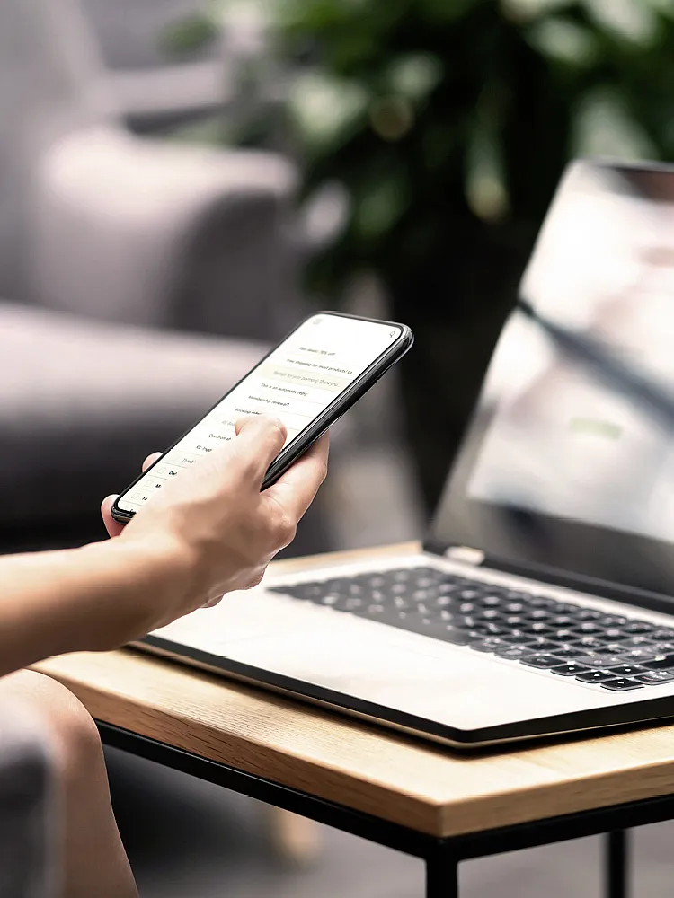 Ein Laptop steht auf einem Couchtisch, davor sitzt eine Frau, die ein Smartphone in der Hand hält