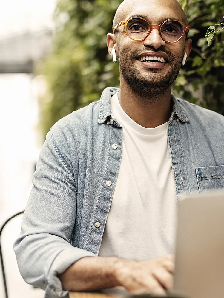 Ein junger Mann mit Brille und Jeanshemd sitzt im freien und arbeitet an seinem Laptop