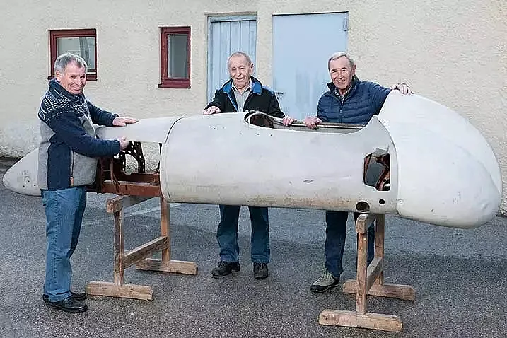 Drei ältere Männer stehen draussen und halten gemeinsam eine unfertige Karosserie eines Oldtimers, die auf Holzgestellen abgestützt ist.