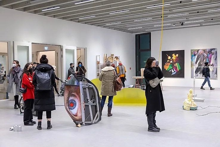 Eine Gruppe von Menschen betrachtet Kunstwerke in einer modernen Galerie. Ein auffälliges Kunstwerk zeigt ein großes Auge auf einem Bildschirm. Die Wände sind mit verschiedenen Gemälden und Installationen dekoriert