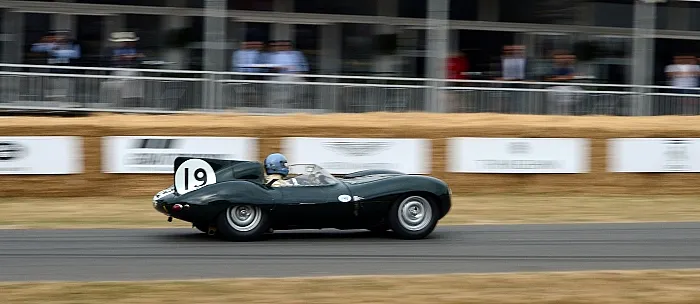 Ein Oldtimer-Rennwagen von Jaguar D-Type mit der Startnummer 19 rast bei einer Rennveranstaltung an verschwommenen Zuschauern vorbei und vermittelt dabei ein Gefühl von Bewegung und Nostalgie.