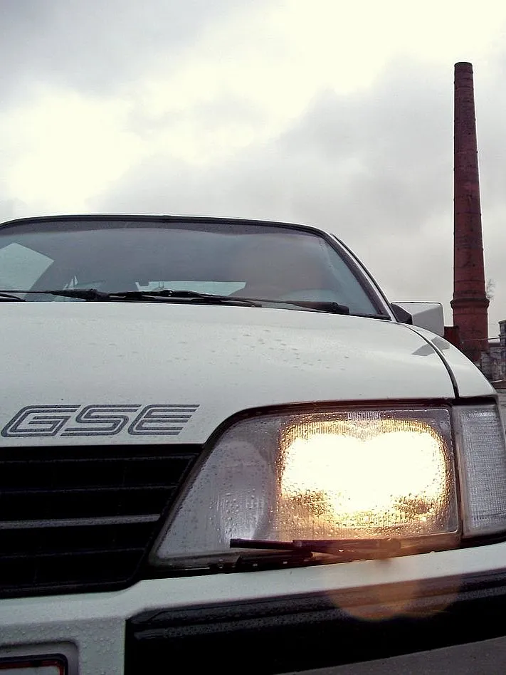 Nahaufnahme der Front eines weißen Autos mit einem GSE-Emblem und eingeschaltetem Scheinwerfer vor dem Hintergrund eines bewölkten Himmels, im Bild ist ein hoher Backsteinschornstein zu sehen.