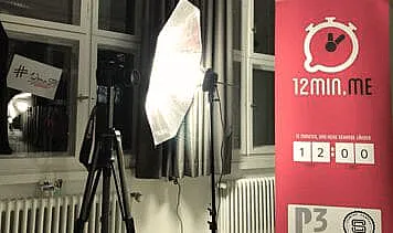 Veranstaltungsaufbau mit rotem Banner von 12min.me, einem Stativ und einer Beleuchtungseinrichtung in einem Raum mit großen Fenstern