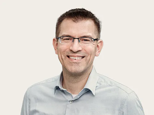 Ein lachender Mann mit Brille und einem hellblauen Hemd, dessen oberer Knop geöffnet ist, vor einem hellen Hintergrundblauen Hemd vor einem neutralen Hintergrund.
