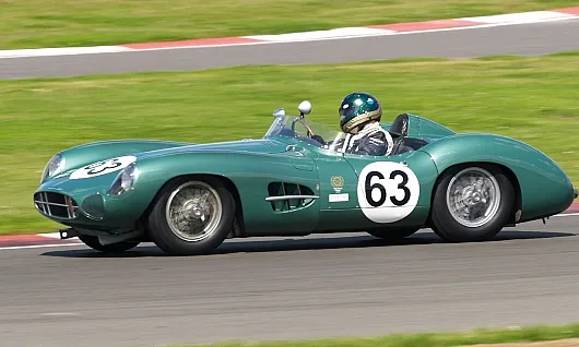 Ein grüner Oldtimer-Rennwagen Aston Martin DBR1 mit der Startnummer 63 rast über die Rennstrecke. Der Fahrer, der einen Helm trägt, beugt sich nach vorne und vermittelt so ein Gefühl von Bewegung und Spannung.
