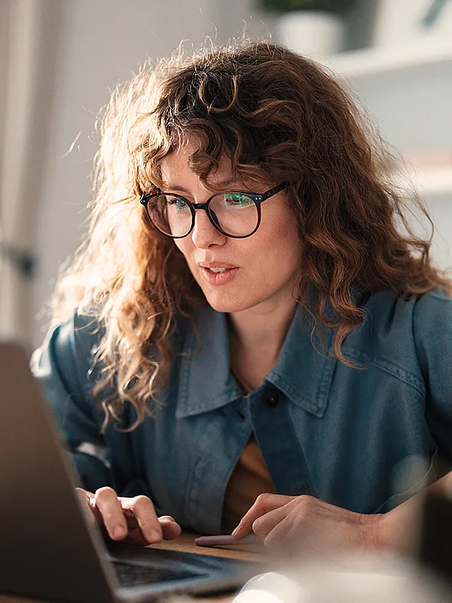 Frau mit Brille und langem brauen lockigen Haar arbeitet konzentriert an einem Laptop.