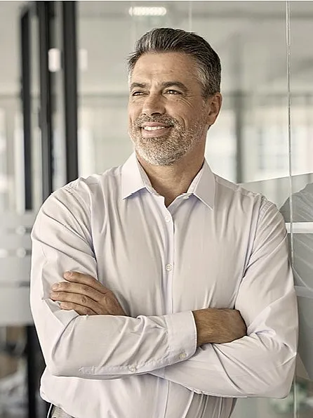 Ein Mann mit grauen Haaren und Bart, der ein helles Hemd trägt, steht lächelnd mit verschränkten Armen in einem modernen Büro.