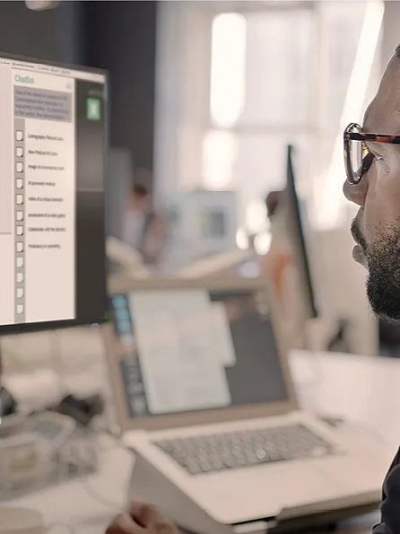 Mann mit schwarzem Hemd und Brille sitzt seitlich vor einem PC-Bildschirm. Im offenen Tab sieht man wie er eine Anfrage in ChatGPT stellt.