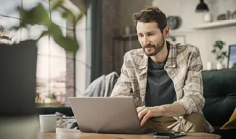 Ein junger Mann mit Bart und Hemd sitzt in einem modernen Zimmer und arbeitet an einem Laptop