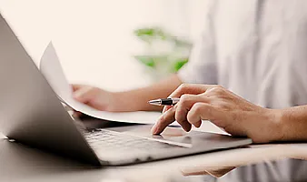 Ein Unbekannter arbeitet am Laptop und hält in einer Hand ein Papier und in der anderen einen Stift