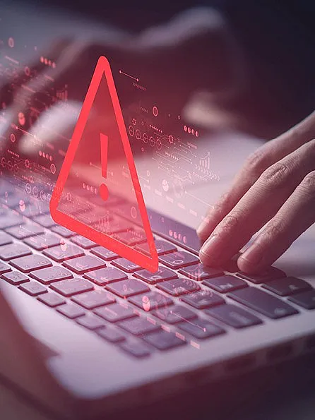 Person sitzt vor einem aufgeklapptem Laptop und tippt mit beiden Händen auf der Tastatur. Ein rotes Warnzeichen erscheint.