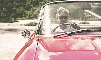 Person fährt ein rotes Cabrio auf einer sonnigen Straße, Blick auf Frontscheibe und Lenkrad.