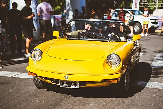 Ein leuchtend gelbes Oldtimer-Cabrio Alfa Romeo Spider mit zwei Insassen fährt während einer Veranstaltung eine Straße entlang. Die Szene ist lebhaft, im Hintergrund stehen Schaulustige.