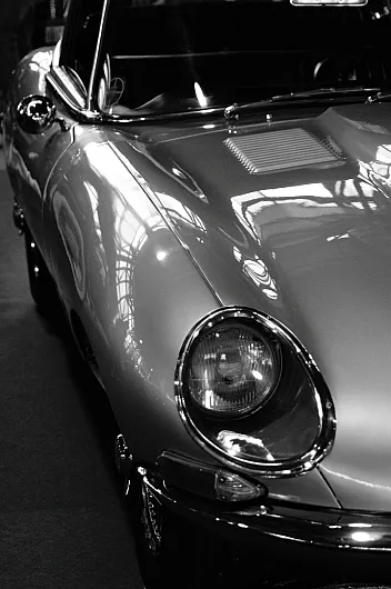 Schwarz-Weiß-Bild eines Oldtimer-Sportwagens Jaguar E-Type mit Fokus auf die vordere rechte Seite. Reflektierende Kurven und runde Scheinwerfer vermitteln einen klassischen, eleganten Stil.