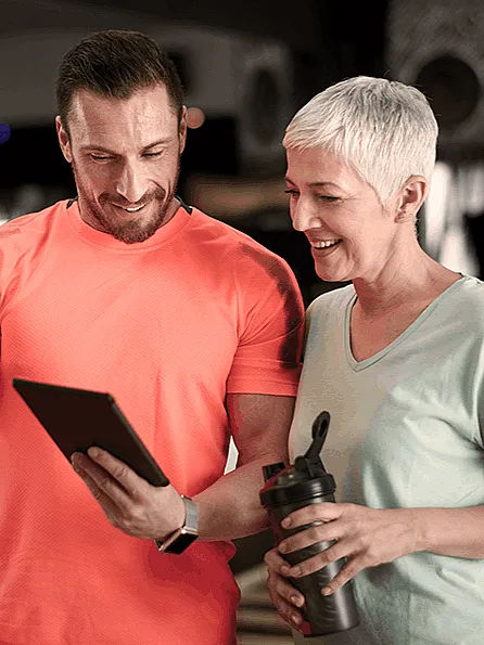 Ein Mann in einem leuchtend orangefarbenen Shirt und eine Frau in einem hellen Shirt stehen in einem Fitnessstudio und lächeln auf ein Tablet. Der Mann feiert mit einer Faustbewegung und drückt damit seine Begeisterung aus.