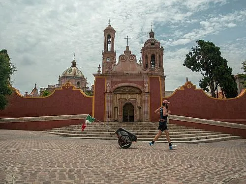 Jonas Deichmann joggt über einen mit Steinen gepflasterten Kirchplatz in Mexico. Die Kirche im Hintergrund ist mit roten Mauern geschützt. Joas zieht einen kleinen Anhänger hinter sich her, an dem eine kleine mexikanische Flagge befestigt ist. Der Himmel ist etwas bewölkt.