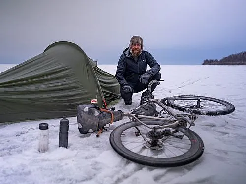 Jonas Deichmann hockt in einer schneebedeckten Landschaft vor einem dunkelgrünen aufgeschlagenen Zelt. Vor ihm liegt sein Fahrrad im Schnee. Jonas trägt dunkle Winterkleidung und eine Fellmütze und lächelt in die Kamera.