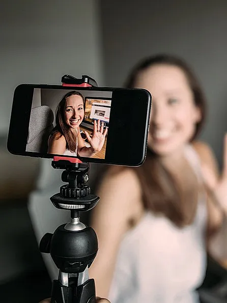Lächelnde Influencerin sitzt vor ihrem PC und hält eine Vlogging-Kamera in der Hand und filmt sich selber