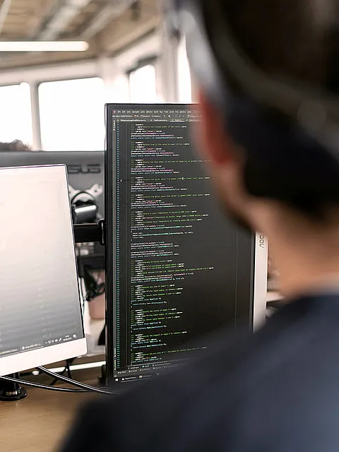 Fotografie zeigt eine Person vor zwei Monitoren mit Programmiercode auf einem Bildschirm, der verschiedene Funktionen und Variablen in farblich hervorgehobener Syntax darstellt. Szene vermittelt Arbeitsumgebung eines Softwareentwicklers mit Fokus auf Codierung und Softwareentwicklung.