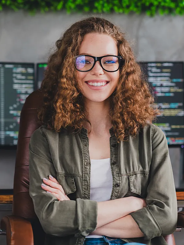 Eine lächelnde Person mit lockigem Haar und Brille sitzt selbstbewusst auf einem Bürostuhl. Hinter ihr zeigen Computerbildschirme eine Programmiersoftware. Hell und konzentriert.