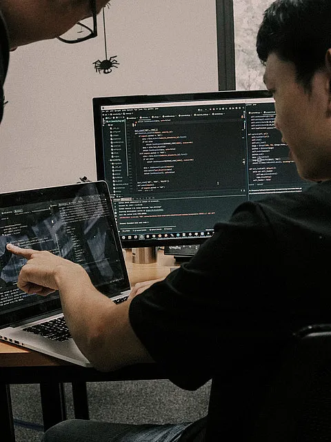 Fotografie zeigt Hände, die auf Laptop-Tastatur tippen, mit zwei Bildschirmen im Hintergrund, die Programmiercode in dunklem Modus anzeigen. Fokus liegt auf Softwareentwicklung, erkennbar an farblich hervorgehobenen Codezeilen und Entwicklerumgebung.