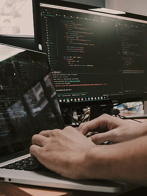 Fotografie zeigt eine Person vor zwei Monitoren mit Programmiercode auf einem Bildschirm, der verschiedene Funktionen und Variablen in farblich hervorgehobener Syntax darstellt. Szene vermittelt Arbeitsumgebung eines Softwareentwicklers mit Fokus auf Codierung und Softwareentwicklung.