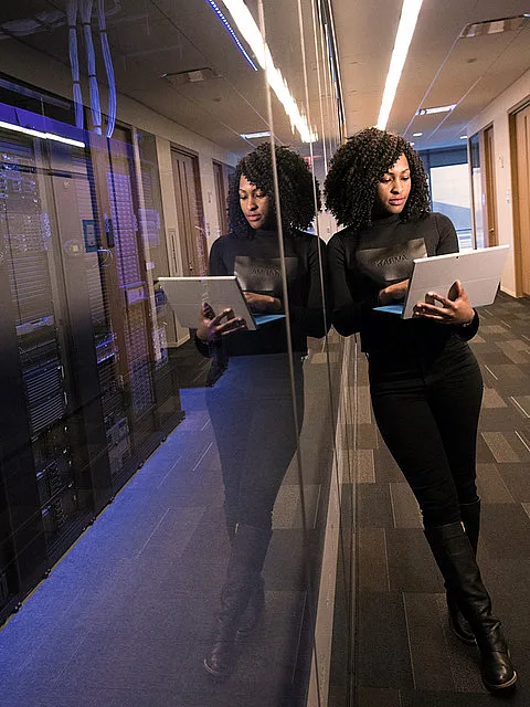 Fotografie einer Frau mit Laptop, die in einem Serverraum oder Rechenzentrum arbeitet, erkennbar an den Serverracks und der technischen Umgebung. Spiegelung an der Glaswand betont moderne Arbeitsumgebung und konzentrierte Tätigkeit im IT-Bereich.