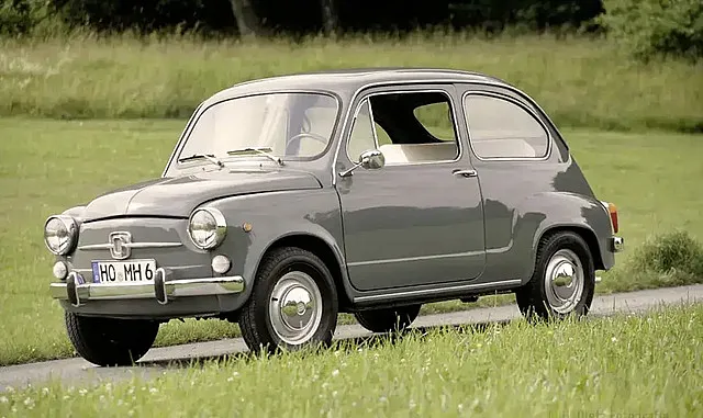 Ein grauer, klassischer Fiat 600, geparkt auf einer Wiese mit Bäumen im Hintergrund. Das Auto hat runde Scheinwerfer, Chromstoßstangen und kleine Räder.