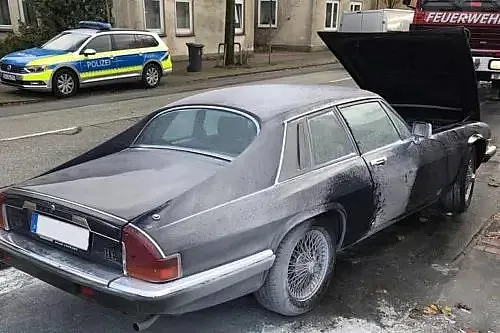 Ein graues Auto mit geöffneter Motorhaube und sichtbaren Schäden am hinteren Teil steht am Straßenrand.