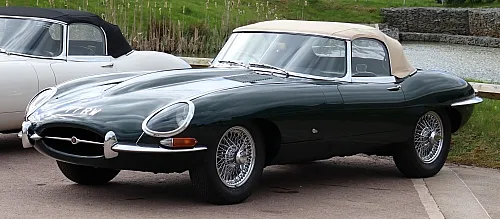 Ein klassisches, dunkelgrünes Jaguar E-Type Cabriolet mit cremefarbenem Verdeck und Speichenrädern, das im Freien neben einem anderen Oldtimer geparkt ist.