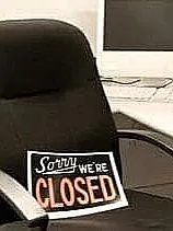 Ein leeres Büro mit einem schwarzen Bürostuhl, auf dem ein Schild mit der Aufschrift "Sorry, WE'RE CLOSED" liegt. Im Hintergrund steht ein alter Computerbildschirm auf einem weißen Schreibtisch, umgeben von verschiedenen Büroartikeln wie Stiften und Papier.