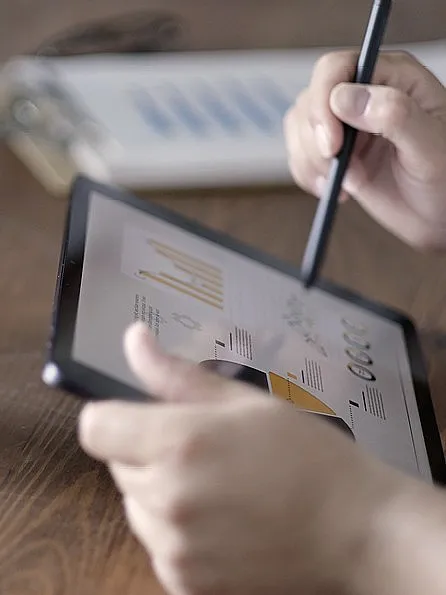 Eine Person arbeitet mit einem Stift auf einem Tablet, auf dem Diagramme angezeigt werden, daneben steht ein kleines Modellhaus auf einem Holztisch. Die Atmosphäre ist konzentriert und analytisch.