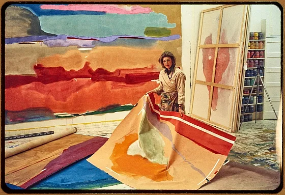 Ein Künstler in einem Atelier mit ausdrucksstarken abstrakten Gemälden, die reich an warmen Farbtönen sind. Der Künstler hält eine große, farbenfrohe Leinwand in der Hand und ist von Malutensilien umgeben, was Kreativität vermittelt.
