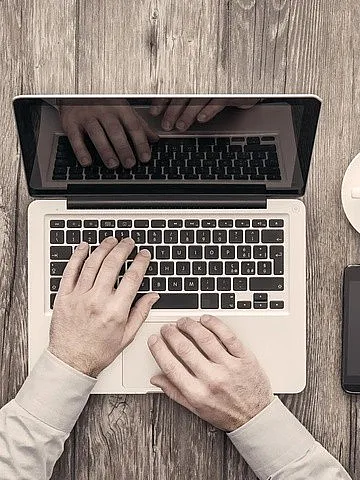 Die Hände einer Person tippen auf einem Laptop. Rechts auf dem Tisch sind eine Kaffeetasse, ein Tablet, ein Smartphone und ein Notizbuch mit einem Stift.