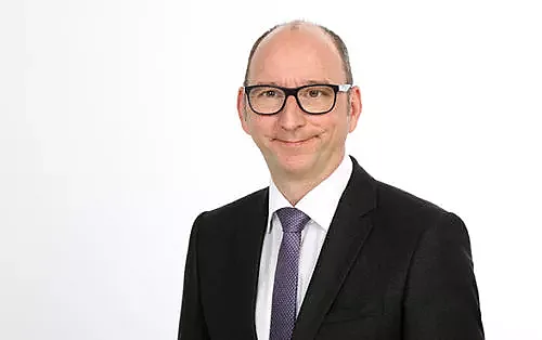 Mario Hartman, vom Management Team, ist in einem schwarzen Anzug mit weißem Hemd und lila Krawatte und steht vor einem weißen Hintergrund.