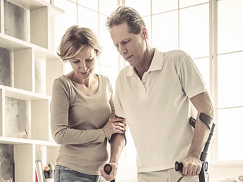 Zwei Personen stehen nah beieinander. Eine Person hält eine Krücke und scheint Unterstützung von der anderen Person zu erhalten, die einen Arm um den Rücken der ersten Person gelegt hat und deren Hand hält. Im Hintergrund ist ein großes Fenster mit natürlichem Licht und einige Regale auf der linken Seite des Bildes zu sehen.