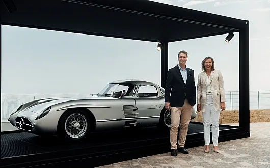 Ein eleganter, silberner Oldtimer, der im Freien in einer Glasvitrine ausgestellt ist. Ein Mann in einem dunklen Blazer und eine Frau in einem hellen Kostüm stehen lächelnd daneben und strahlen Stolz aus.