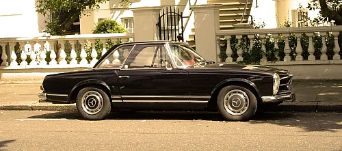 Ein schwarzes Mercedes-Benz-Coupé im Vintage-Stil, das auf einer sonnenbeschienenen Stadtstraße geparkt ist, hinter dem sich kunstvoll verzierte weiße Geländer und Treppen eines Stadthauses befinden.