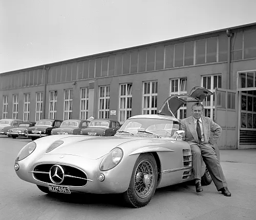 Ein Mann im Anzug lehnt an einem Mercedes-Benz 300SL im Retro-Stil mit geöffneten Flügeltüren. Die Szene spielt sich vor einem Industriegebäude mit geparkten Autos ab.