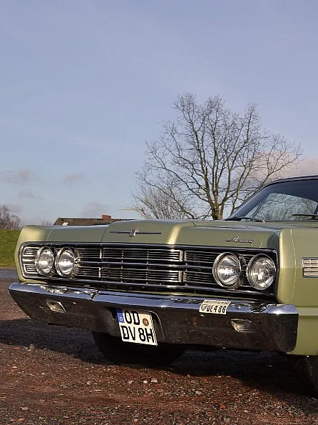 Ein klassischer grüner Ford Galaxie 500 ist auf einer Kiesfläche geparkt. Der Himmel ist klar und im Hintergrund sind einige Bäume zu sehen. Das Auto hat Chromdetails, runde Scheinwerfer und ein schwarzes Dach.