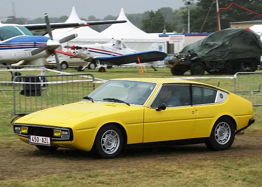 Ein gelber Oldtimer-Sportwagen Simca Bagheera mit schlankem Design fährt auf Gras in der Nähe eines kleinen Flugzeugs und einiger Zelte. Die Szene vermittelt eine nostalgische, klassische Atmosphäre.