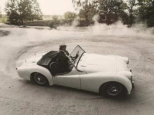 Ein weißes Cabrio driftet auf einer Schotterfläche und wirbelt Staub auf. Im Hintergrund sind Bäume und Grünflächen zu sehen.