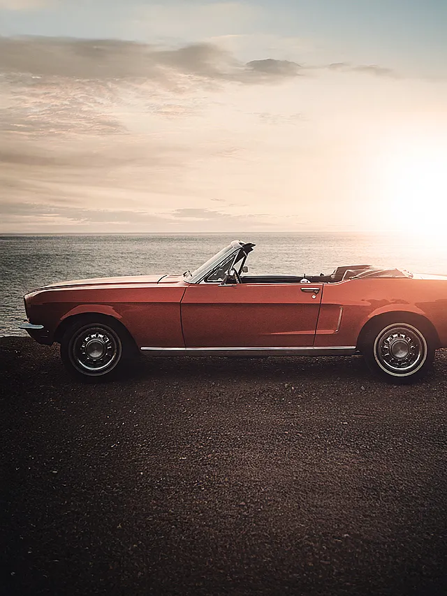 Roter Oldtimer-Cabrio steht seitlich auf einer Küstenstraße mit Blick auf das Meer bei Sonnenuntergang.